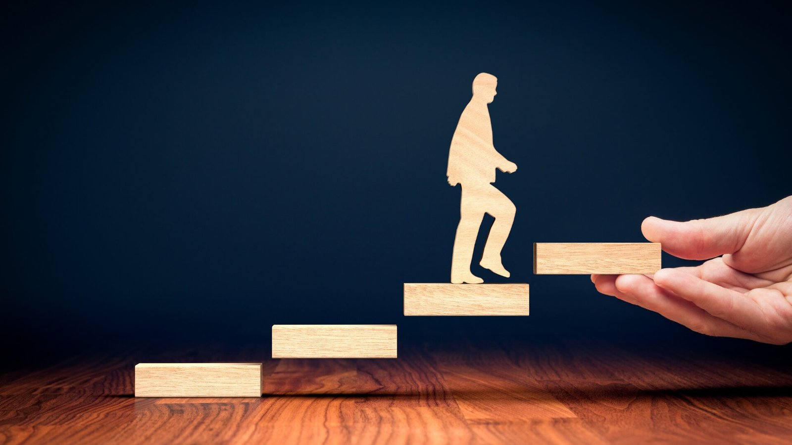 Kişisel gelişimi simgeleyen ahşap bir adam figürü, bir el tarafından desteklenen merdiven basamaklarında yukarı doğru ilerliyor.